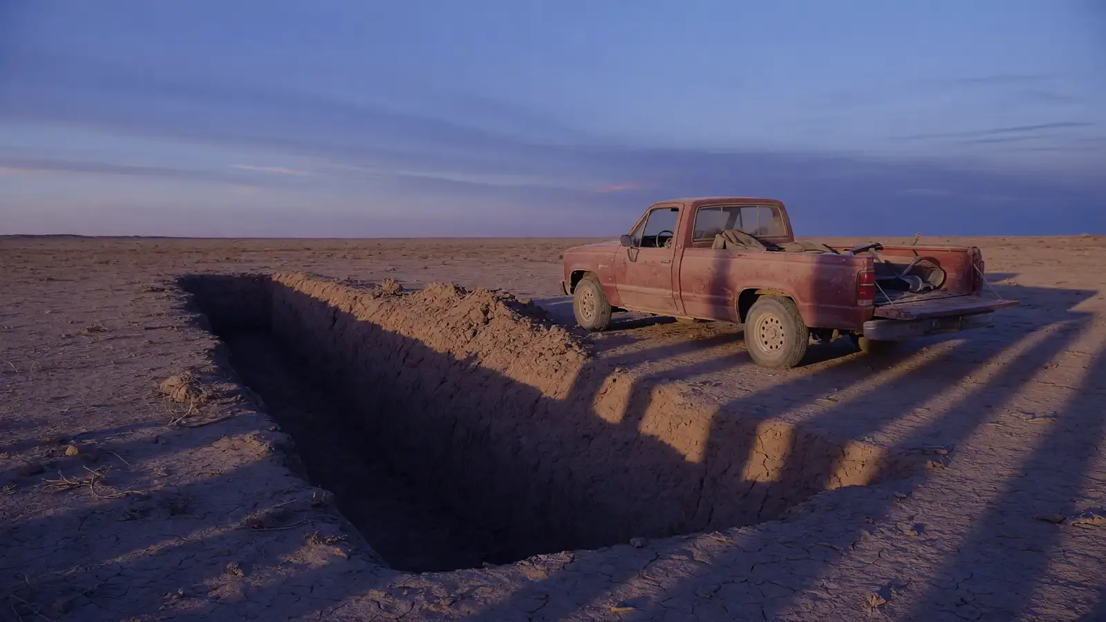 Empty desert excavation site at dusk with a parked pickup and a dark trench, no people or readable signs.
