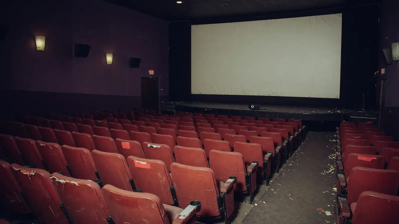 An empty mid-size movie theater with worn red seats and a blank white screen.